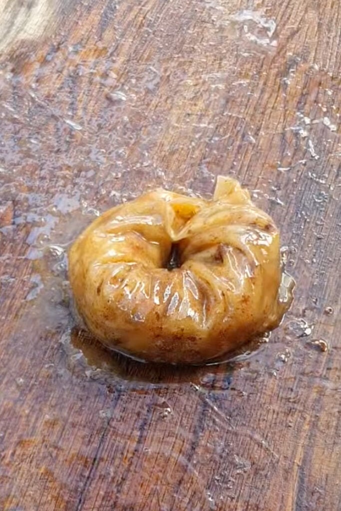 A round rice paper apple donut with a glossy, brown exterior sits on a wet wooden surface, glistening with oil or sauce.