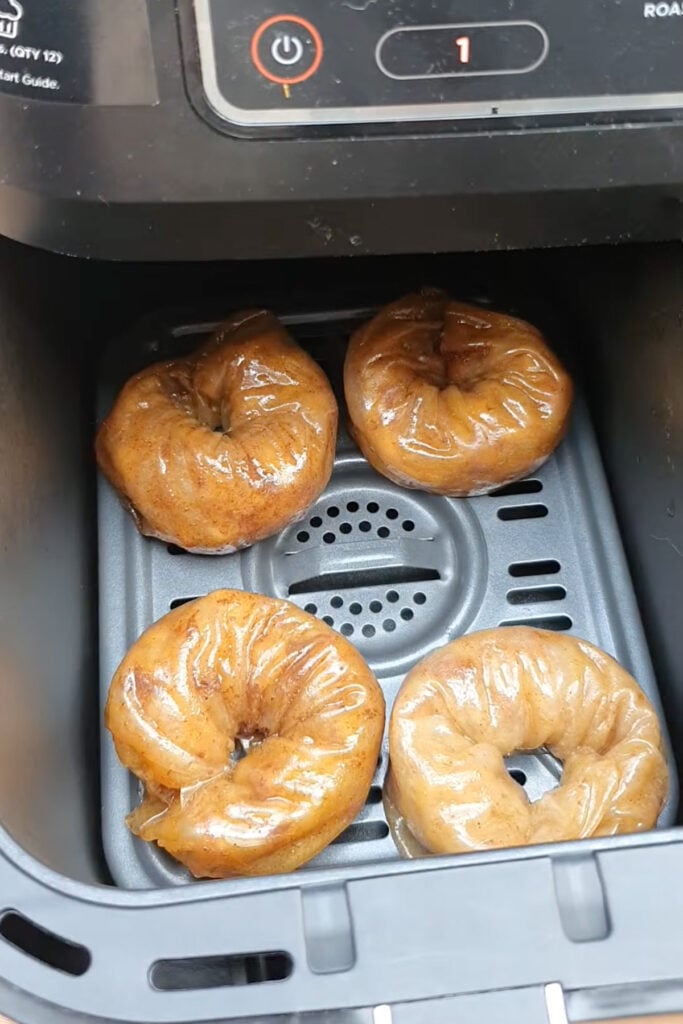 Four rice paper apple donuts are placed inside an air fryer basket, ready to be cooked. The air fryer control panel is visible at the top of the image.