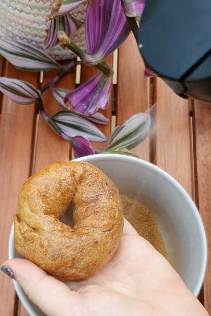 A hand holds an air fryer apple donut over a bowl of cinnamon sugar as they spray oil on it, with purple flowers and a wooden table in the background.