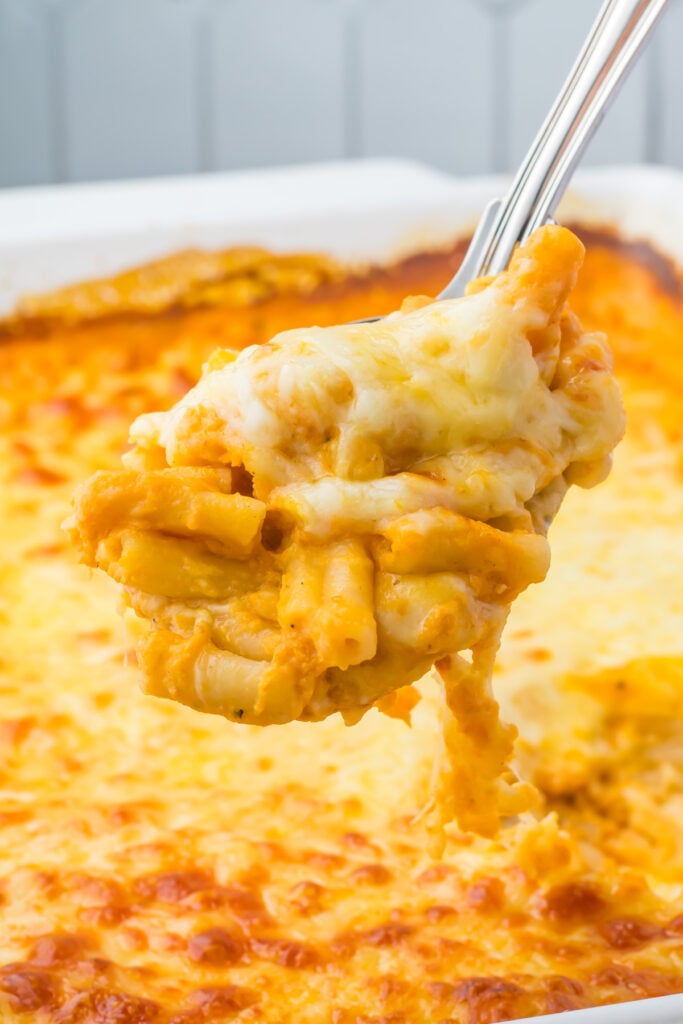 A close-up of a metal serving spoon lifting a portion of baked pumpkin mac and cheese from a casserole dish.