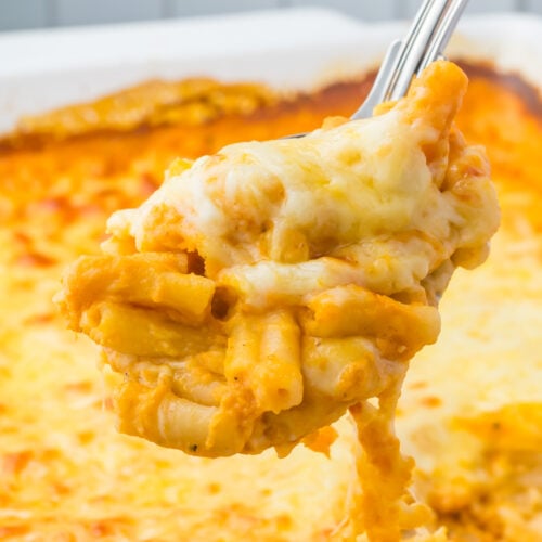 A close-up of a serving spoon lifting cheesy baked pumpkin mac and cheese with a golden, melted cheese topping from a casserole dish.