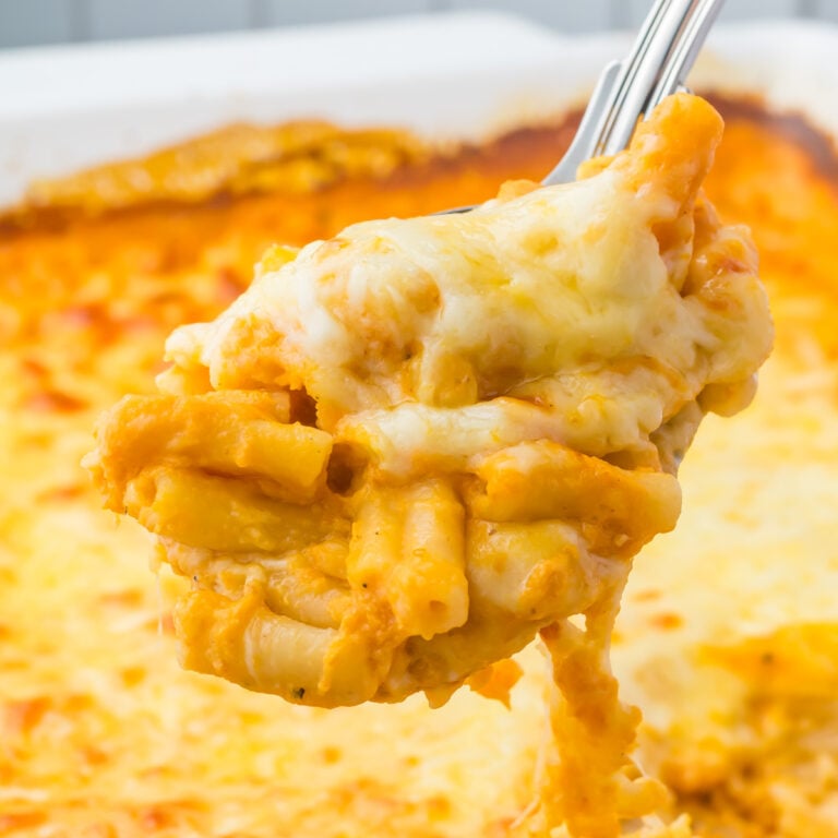 A close-up of a serving spoon lifting cheesy baked pumpkin mac and cheese with a golden, melted cheese topping from a casserole dish.