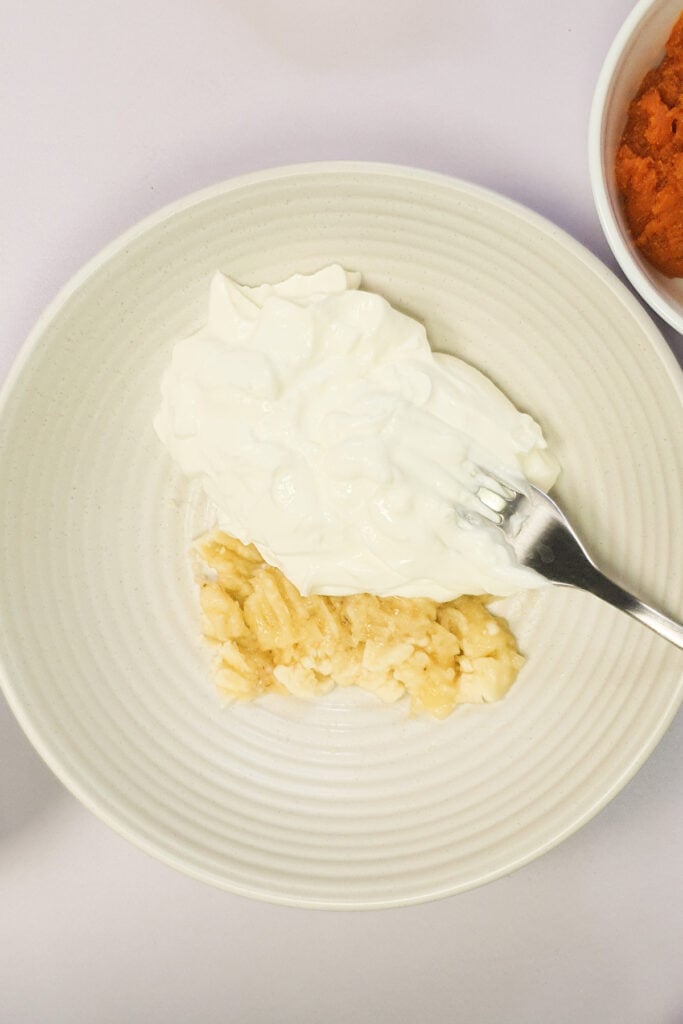 A plate with mashed banana and a serving of plain yogurt, with a fork resting on the mashed banana.