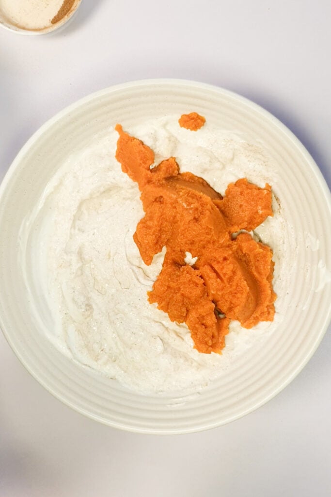 A white bowl contains a mixture of yogurt and orange pumpkin puree on a white surface.