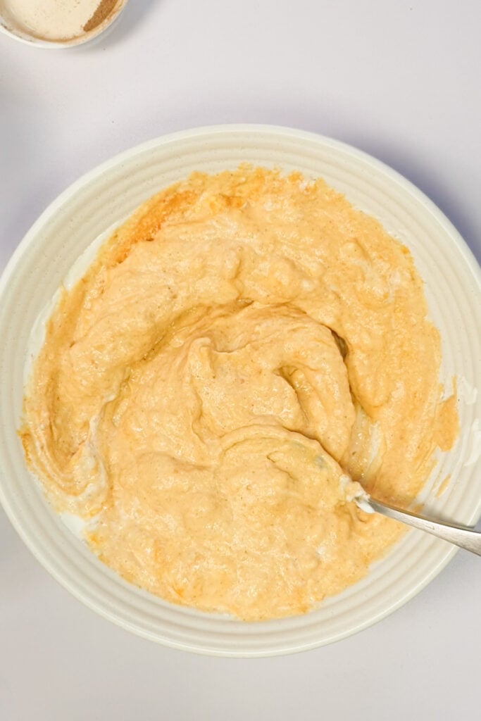 A white bowl containing a creamy, light orange mixture with a spoon resting inside, placed on a white surface.