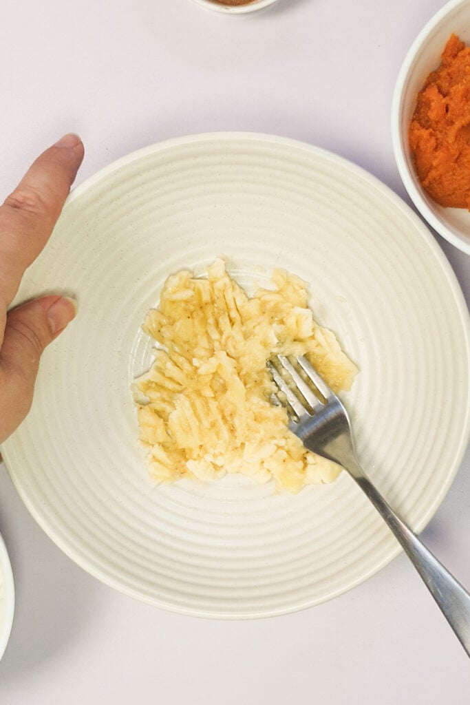 A hand holds a bowl with mashed banana and a fork on it. Bowls with other ingredients are partially visible around the plate.