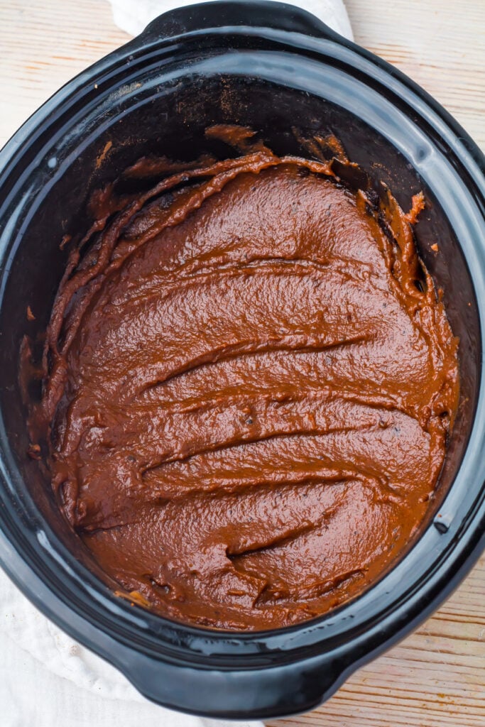 Thick, smooth brown apple butter spread evenly across the bottom of a black slow cooker, with visible swirl marks on the surface.