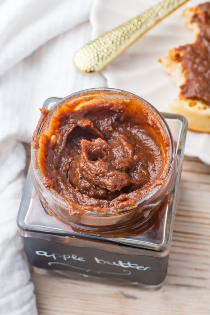 Open jar of slow cooker apple butter on a wooden surface, with a knife and a partially spread piece of bread visible in the background.