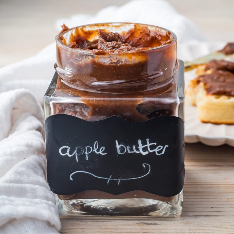 A square glass jar labeled “apple butter” filled with a thick, dark brown spread sits on a light wooden surface near a white cloth.