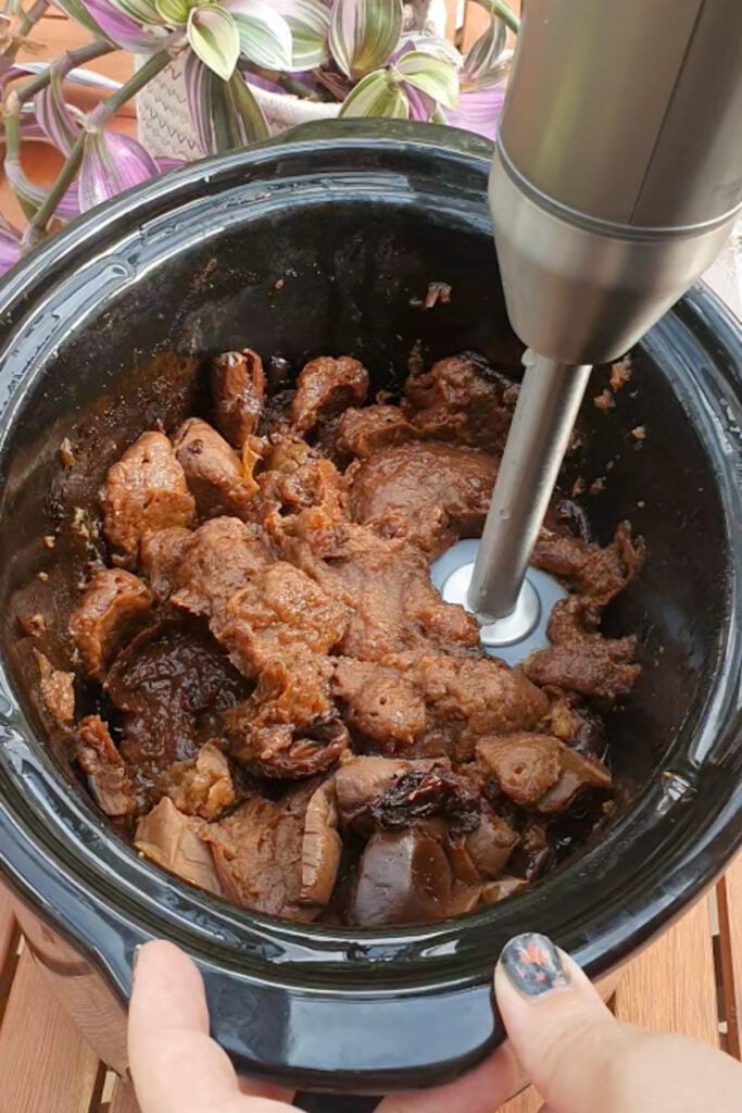 A hand holds a stick blender in a slow cooker filled with cooked apples, preparing to blend the contents.