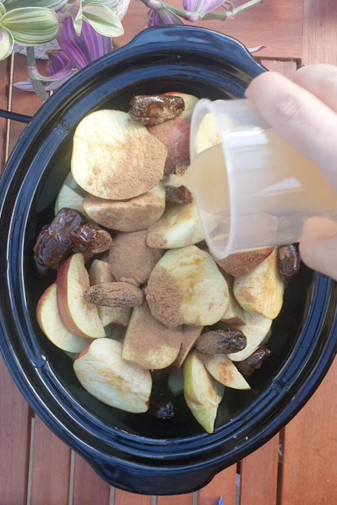 A hand pours liquid into a slow cooker filled with sliced apples, dates, and brown spices on a wooden table with plants nearby.