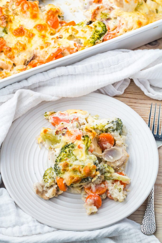 A serving of chicken, broccoli, and rice casserole topped with melted cheese on a white plate next to a fork and a baking dish.