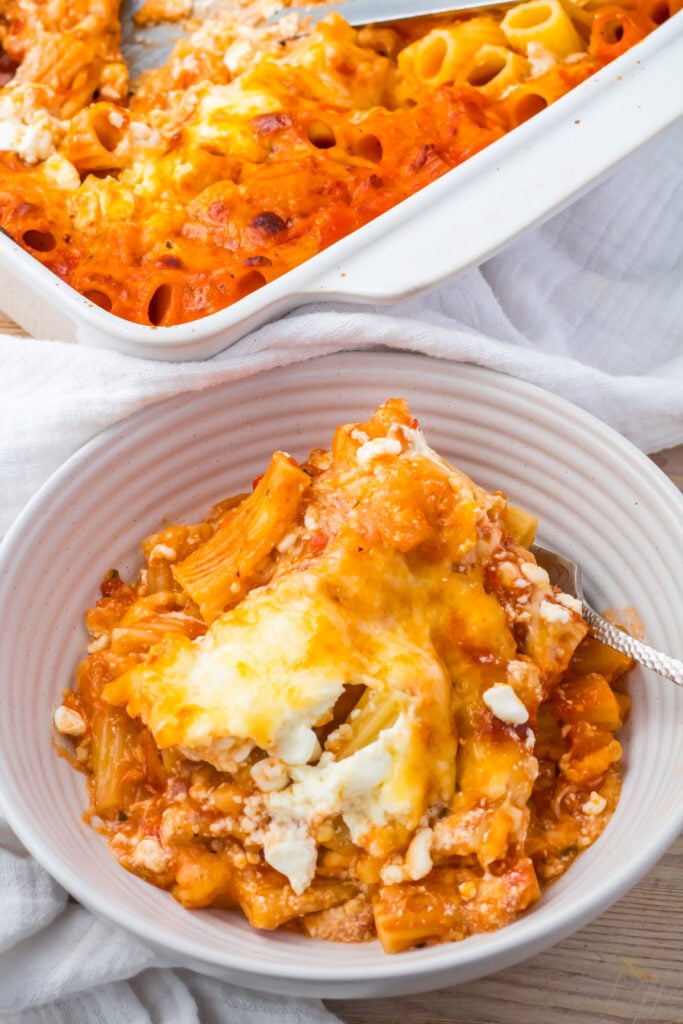 A serving of cottage cheese baked ziti with melted cheese in a white bowl, with a baking dish of the same pasta in the background.