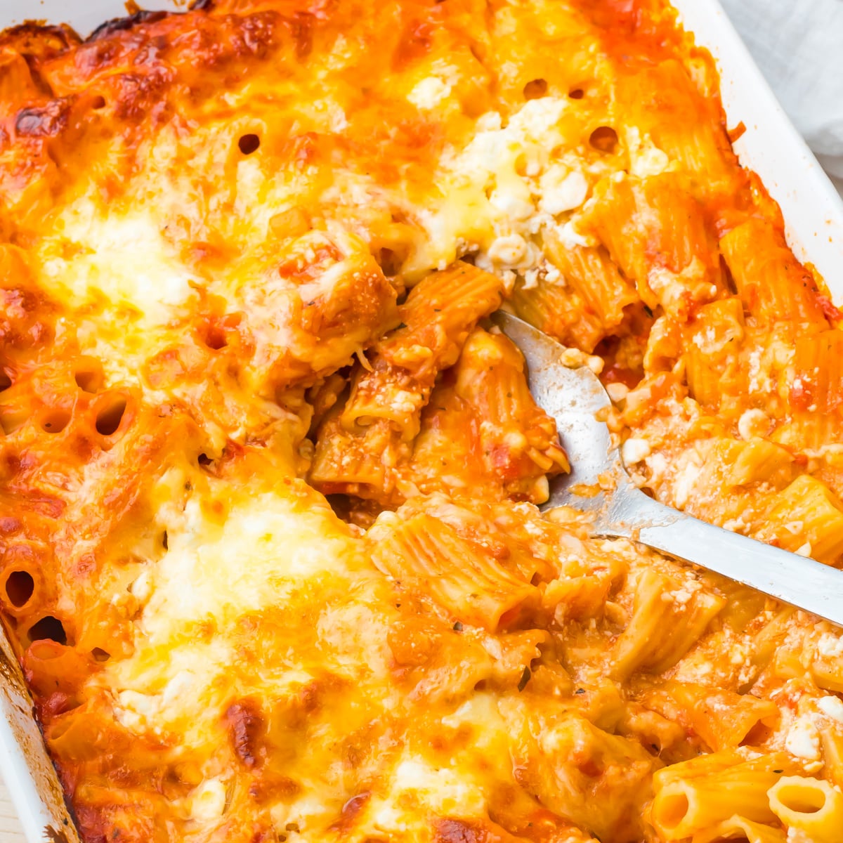 Cottage cheese baked ziti pasta with melted cheese and tomato sauce in a white casserole dish, being served with a metal spoon.