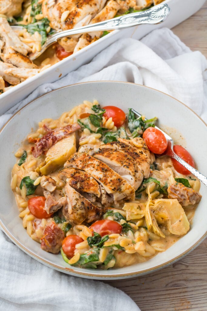 A bowl of marry me chicken orzo pasta with sliced chicken, spinach, cherry tomatoes, and artichokes, with a portion of the same dish in a baking dish nearby.
