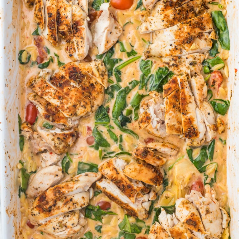 an overhead photo of marry me chicken orzo bake: sliced seasoned chicken breasts served over a creamy orzo pasta bake with spinach and cherry tomatoes in a white baking dish.