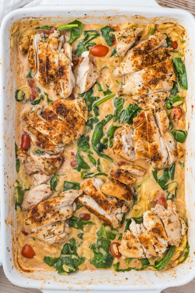 an overhead photo of marry me chicken orzo bake: sliced seasoned chicken breasts served over a creamy orzo pasta bake with spinach and cherry tomatoes in a white baking dish.