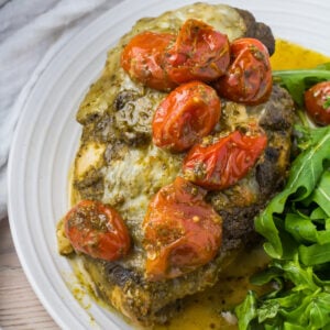 Pesto chicken bake topped with melted cheese and roasted cherry tomatoes, served with a side of arugula on a white plate.