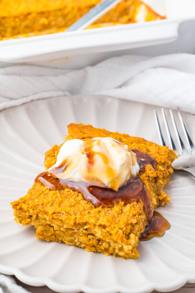 A square piece of pumpkin baked oatmeal topped with whipped cream and syrup sits on a white plate with a fork beside it.