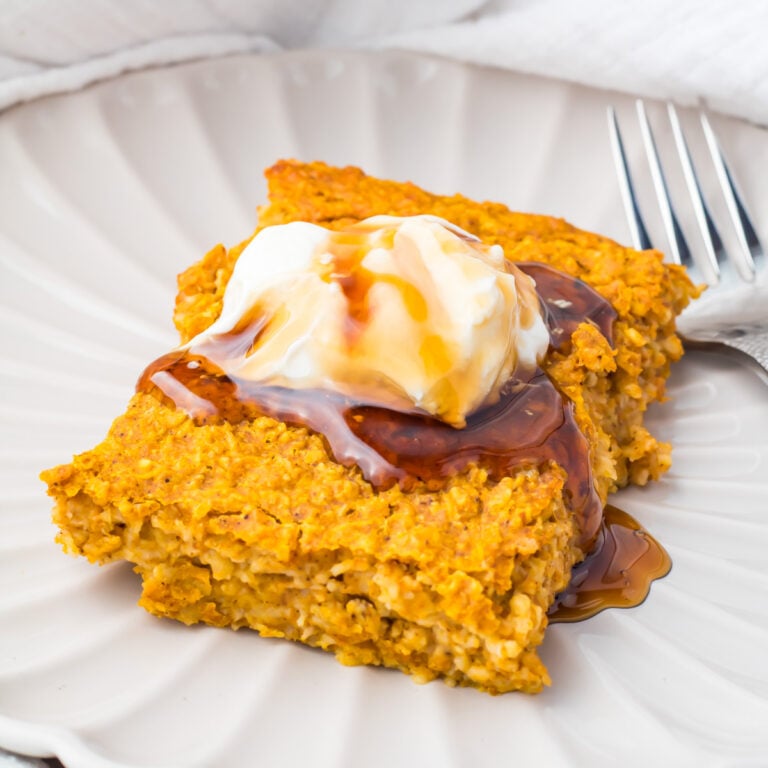 A square piece of pumpkin baked oatmeal topped with a dollop of yogurt and a drizzle of syrup, served on a white plate with a fork.