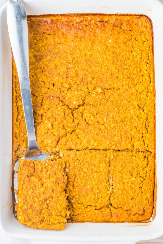 A pumpkin baked oatmeal dish with a golden-brown top, cut into squares, with a metal spatula lifting one piece in a white baking pan.