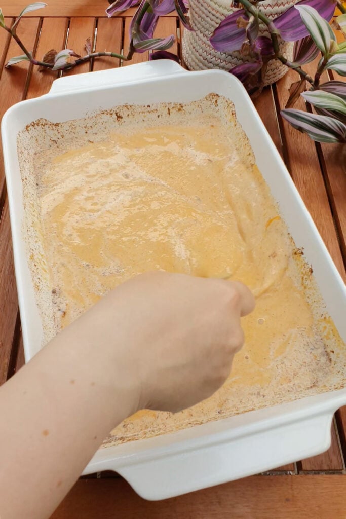 A hand stirs a creamy orange mixture in a rectangular white baking dish on a wooden table with a potted plant nearby.