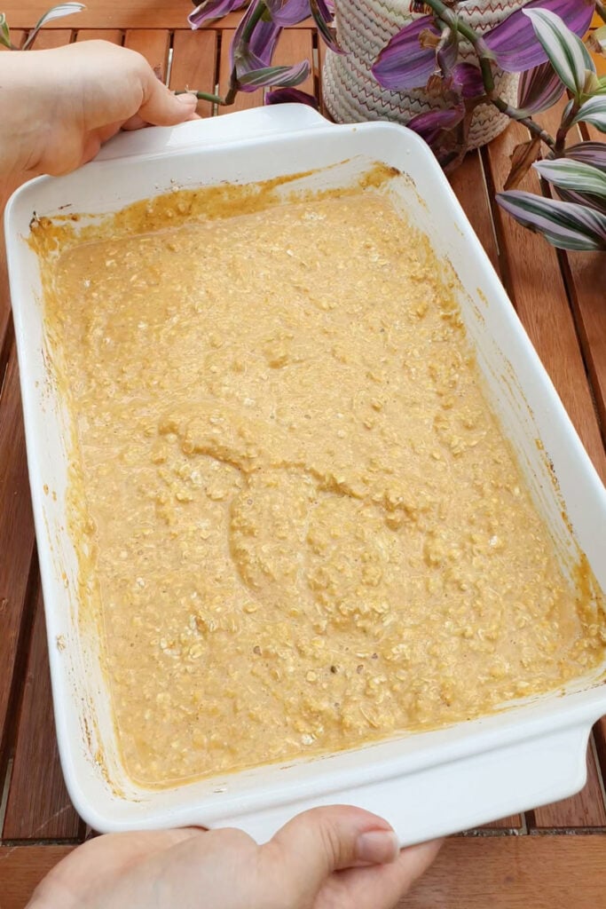 A person holds a white baking dish filled with an unbaked mixture of oats and batter on a wooden table with a potted plant nearby.