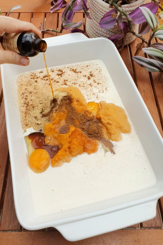 A hand pours vanilla into a white dish containing wet ingredients on a wooden table near a potted plant.