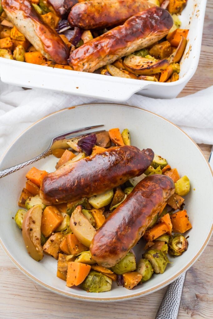 Sausage sweet potato bake served in a bowl with roasted vegetables including Brussels sprouts, sweet potatoes, and apples, with a baking dish in the background.